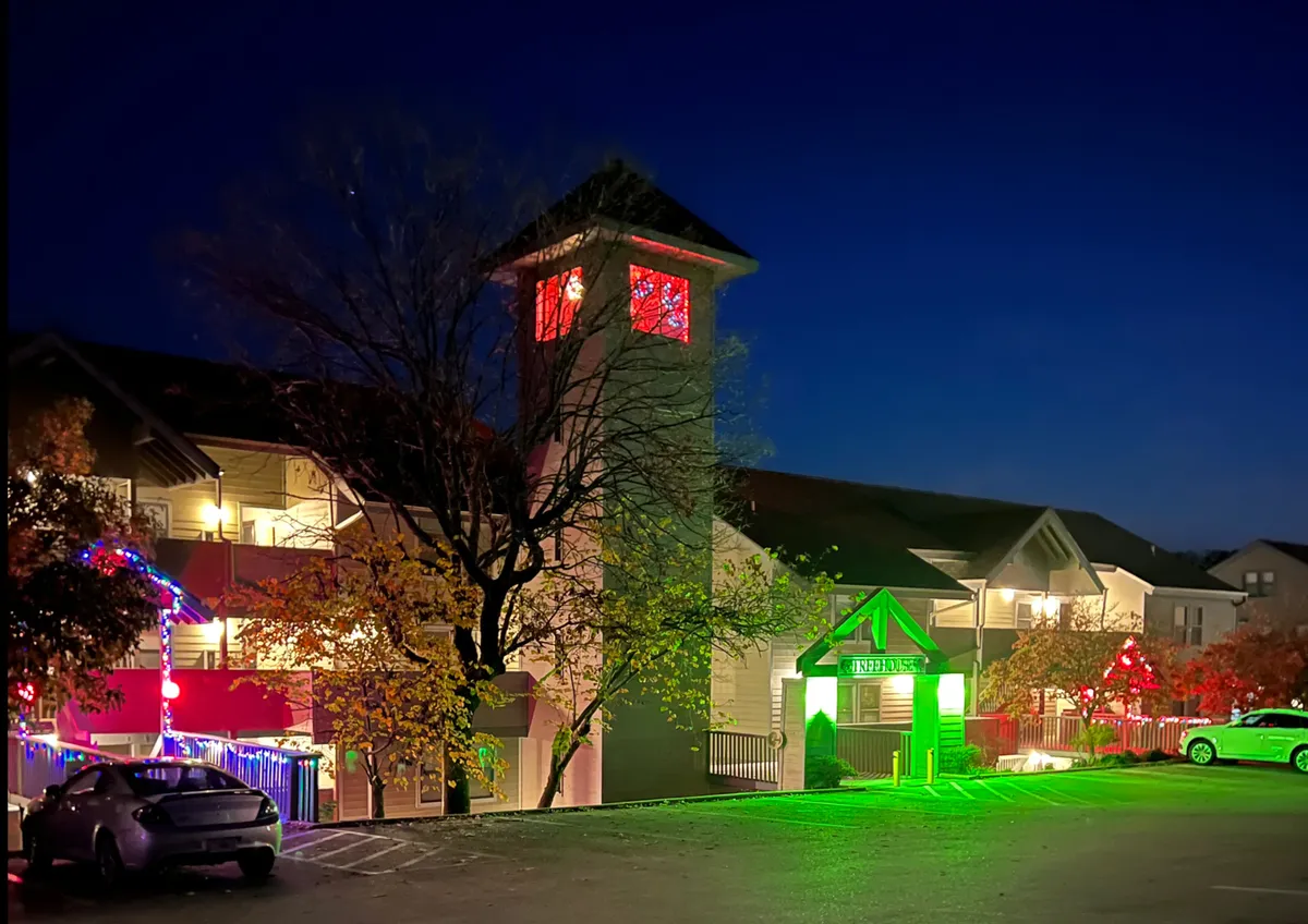 An exterior nighttime scene glows with decorative holiday lights, featuring a tower with red-lit windows and a warmly illuminated green entrance. The festive ambiance suggests winter celebrations and community events, capturing the sense of holiday spirit and togetherness.