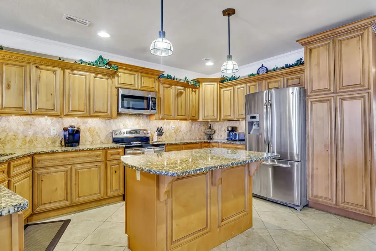 A spacious, modern kitchen featuring gleaming granite countertops, an expansive central island with pendant lighting, and high-end stainless steel appliances. Warm wooden cabinetry lines the walls, and the setup invites gatherings for cooking and entertaining. Natural light pours in, creating an inviting atmosphere for culinary activities.